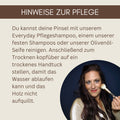 Frau mit langen braunen Haaren hält einen veganen Make-up-Pinsel von Thats me Organic® vor ihrem Gesicht und lächelt vor dunkelblauem Hintergrund.