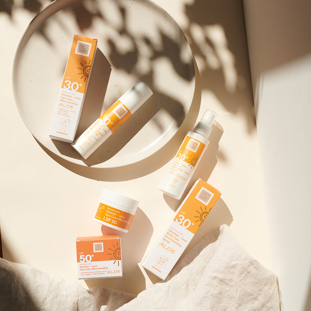 Vier Thats me Organic® NATRUE-zertifizierte Sonnenschutzprodukte in orangefarbener und weißer Verpackung sind auf einer beigen Platte mit einem Stoffhintergrund abgebildet.