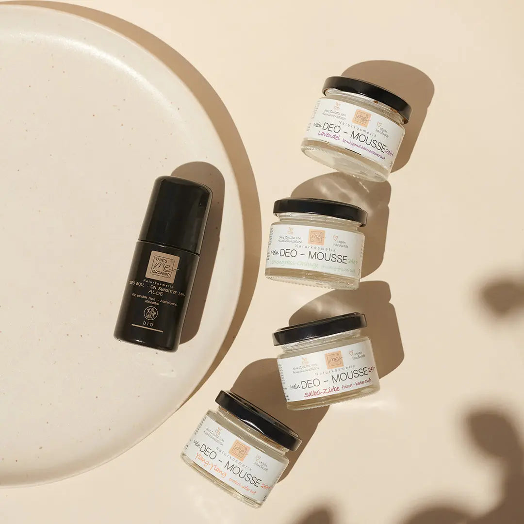 Vier Glastiegel und ein schwarzer Flakon von Thats me Organic® Bio-Deo stehen auf einer beigen Fläche neben einem runden Teller und zeigen die raffinierte Eleganz von natürlichem Deo für Ihren Alltag.