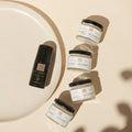 Vier Glastiegel und ein schwarzer Flakon von Thats me Organic® Bio-Deo stehen auf einer beigen Fläche neben einem runden Teller und zeigen die raffinierte Eleganz von natürlichem Deo für Ihren Alltag.