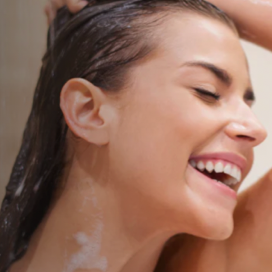 Lächelnde Frau mit geschlossenen Augen, die sich unter der Dusche die Haare wäscht, wobei Wasser und Seife in ihren Haaren zu sehen sind.