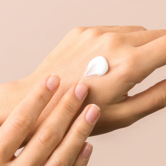 Eine Hand mit einem Klecks weißer Creme, die von einer anderen Hand vor einem beigen Hintergrund aufgetragen wird.