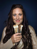 Eine lächelnde Frau mit langen braunen Haaren hält vier vegane Thats me Organic® Make-up-Pinsel vor einem dunklen Hintergrund.
