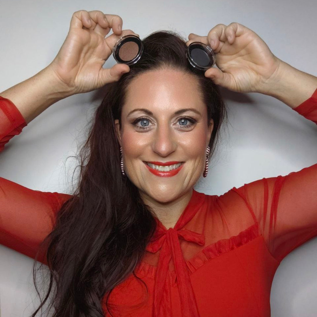 Frau in rotem Oberteil lächelt und hält zwei runde Thats me Organic® Naturkosmetik Lidschatten-Dosen über ihrem Kopf vor weißem Hintergrund.