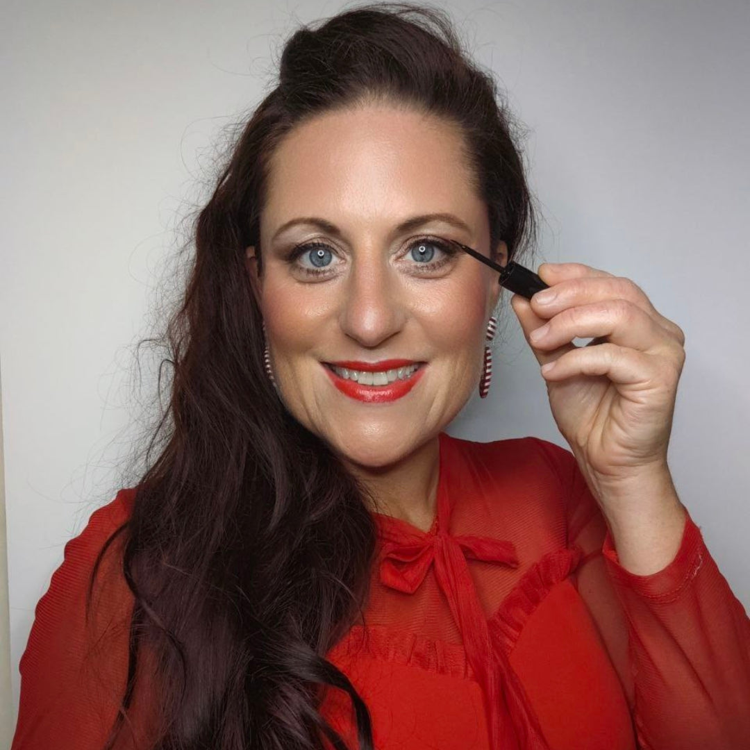 Eine Frau in einem roten Oberteil lächelt, während sie die Thats me Organic® Bio Eyeliner Wimperntusche auf ihre empfindlichen Augen aufträgt. Der Hintergrund ist einfarbig und völlig frei von Titandioxid.