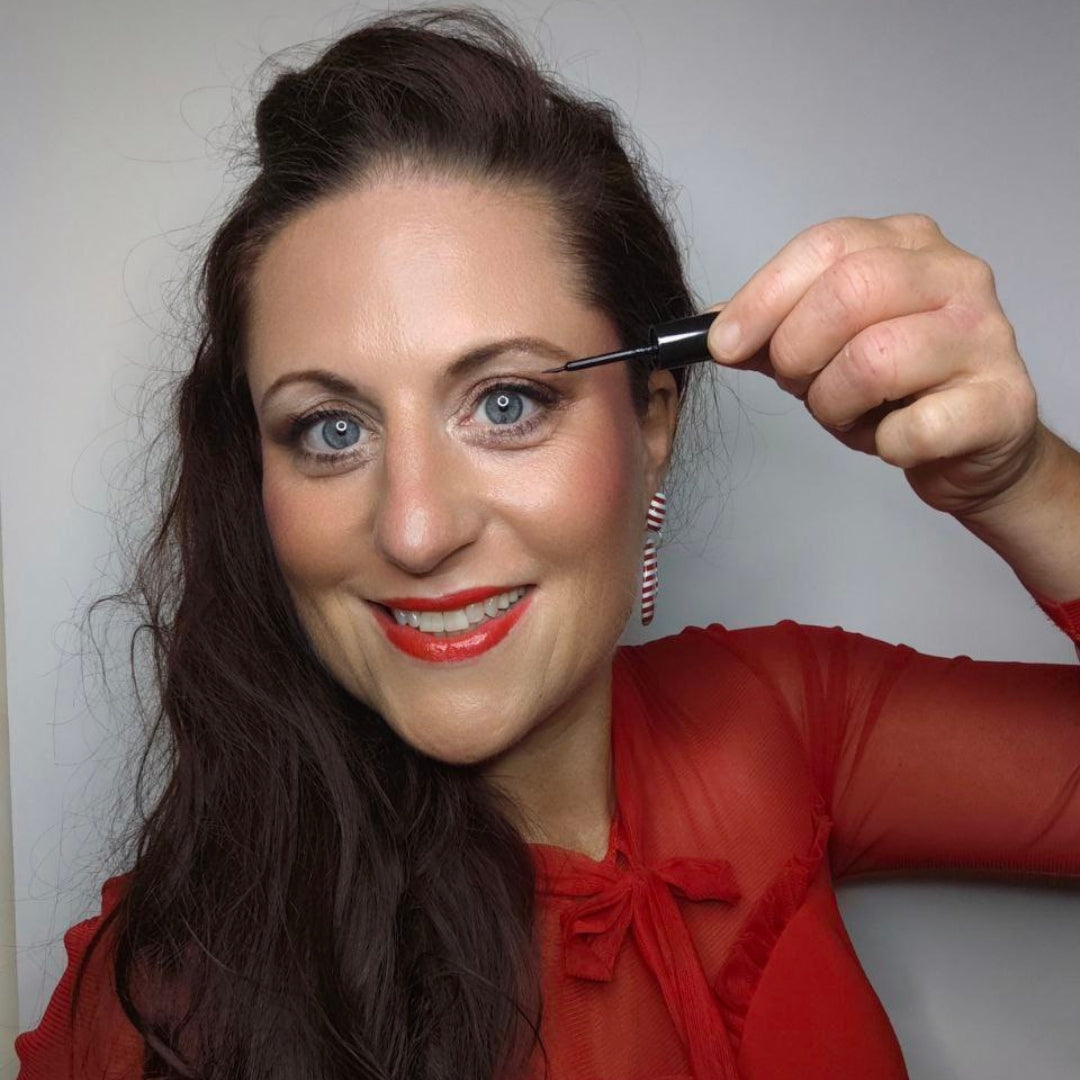 Eine Frau mit rotem Oberteil und rotem Lippenstift lächelt, während sie Thats me Organic® Bio Eyeliner, ideal für empfindliche Augen, vor einem schlichten Hintergrund aufträgt.