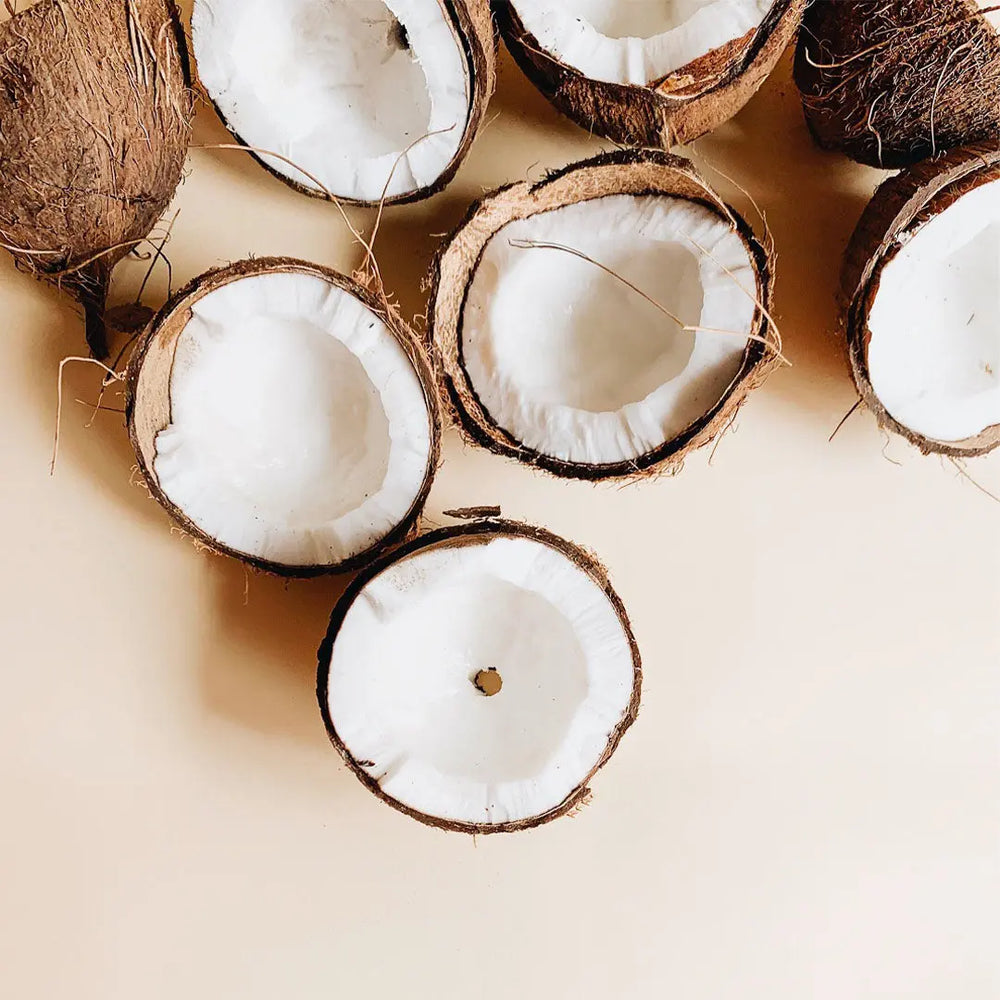 Halbierte Kokosnüsse mit weißem Fruchtfleisch und braunen Schalen auf hellbeiger Oberfläche inspirieren Thats me Organic® festes Shampoo - ideal für empfindliches Haar.