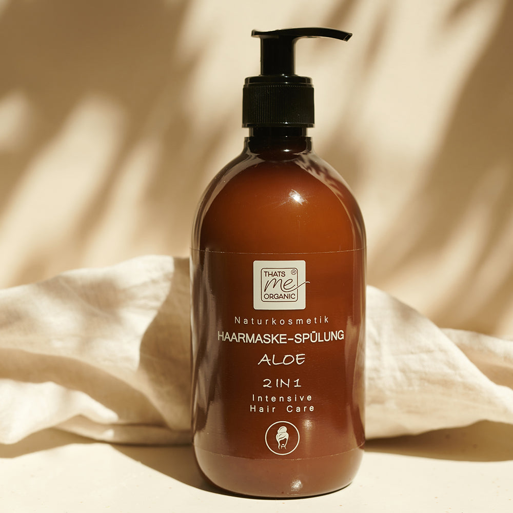 Eine braune Pumpflasche mit Thats me organic® aloe 2-in-1 intensive Bio-Haarmaske und Spülung für natürliche Haarpflege steht auf einem beigen Stoffhintergrund.